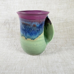 Puede incluir: Una taza de cerámica hecha a mano con un diseño único. La taza presenta un borde morado, que pasa a tonos azules y verdes en el cuerpo. El asa es de color verde claro. Una hermosa pieza de arte funcional.