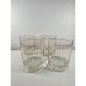 May include: Set of four clear drinking glasses with gold vertical stripes. The glasses have a rounded shape and a gold band around the rim. The glasses are likely for serving beverages.