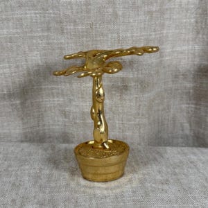 May include: A gold-colored metal sculpture of a small tree planted in a round pot. The tree features a smooth, vertical trunk and a wide, branching canopy with rounded, leaf-like protrusions. The entire object has a polished, reflective finish.