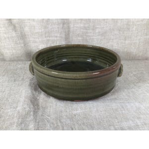 Puede incluir: Un cuenco de cerámica de color verde oliva con acabado brillante y textura acanalada horizontal en las paredes exteriores. El cuenco cuenta con dos pequeñas asas integradas y un interior oscuro y liso con un borde de color marrón rojizo.