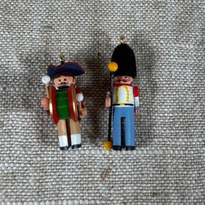 May include: Two wooden ornaments featuring painted figures. One figure wears a blue hat and holds a green and red drum with white mallets. The other figure wears a tall black fuzzy hat, a white jacket with yellow trim, and blue pants, holding a staff.