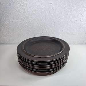 May include: A stack of round ceramic plates featuring a dark brown finish with a subtle speckled texture. Each plate has a slightly raised rim and a smooth, matte-like surface, creating a uniform appearance throughout the entire set of dinnerware.