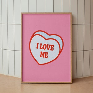 A framed retro-inspired poster featuring two overlapping blue-colored candy hearts with the words 'I Love Me' in bold red text. The design is set against a soft pink background. Displayed against a tiled wall.
