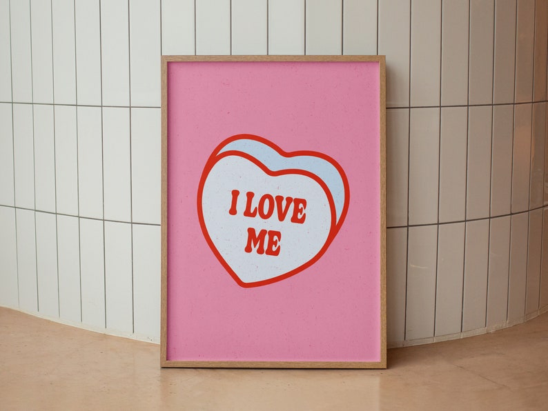 A framed retro-inspired poster featuring two overlapping blue-colored candy hearts with the words 'I Love Me' in bold red text. The design is set against a soft pink background. Displayed against a tiled wall.