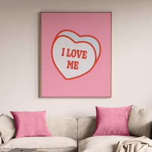 A framed retro-inspired poster featuring two overlapping blue-colored candy hearts with the words 'I Love Me' in bold red text. The design is set against a soft pink background. Displayed above a trendy cream couch with pink cusions.