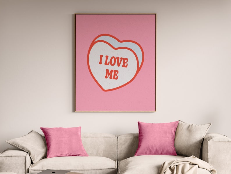 A framed retro-inspired poster featuring two overlapping blue-colored candy hearts with the words 'I Love Me' in bold red text. The design is set against a soft pink background. Displayed above a trendy cream couch with pink cusions.
