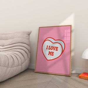 A framed retro-inspired poster featuring two overlapping blue-colored candy hearts with the words 'I Love Me' in bold red text. The design is set against a soft pink background. Displayed in a trendy room.