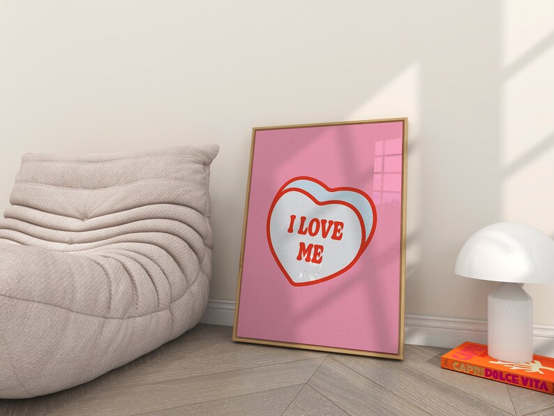 A framed retro-inspired poster featuring two overlapping blue-colored candy hearts with the words 'I Love Me' in bold red text. The design is set against a soft pink background. Displayed in a trendy room.