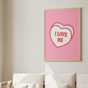 A framed retro-inspired poster featuring two overlapping blue-colored candy hearts with the words 'I Love Me' in bold red text. The design is set against a soft pink background. Displayed against a trendy couch.