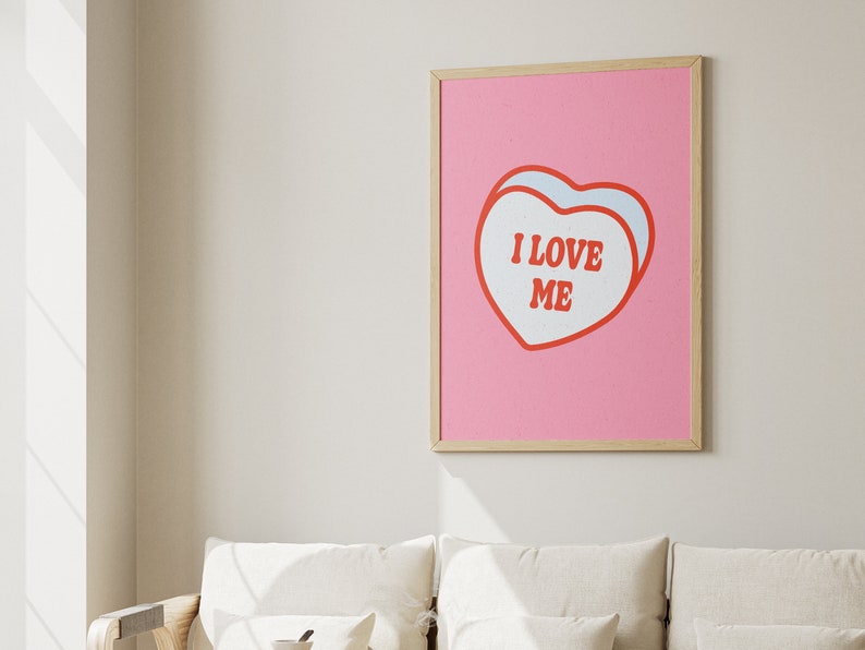 A framed retro-inspired poster featuring two overlapping blue-colored candy hearts with the words 'I Love Me' in bold red text. The design is set against a soft pink background. Displayed against a trendy couch.