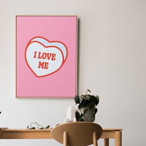 A framed retro-inspired poster featuring two overlapping blue-colored candy hearts with the words 'I Love Me' in bold red text. The design is set against a soft pink background. Displayed above a dining table.