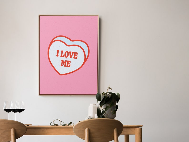 A framed retro-inspired poster featuring two overlapping blue-colored candy hearts with the words 'I Love Me' in bold red text. The design is set against a soft pink background. Displayed above a dining table.