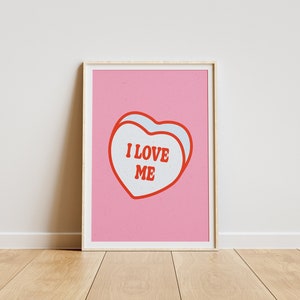 A framed retro-inspired poster featuring two overlapping blue-colored candy hearts with the words 'I Love Me' in bold red text. The design is set against a soft pink background. Displayed against a white wall.