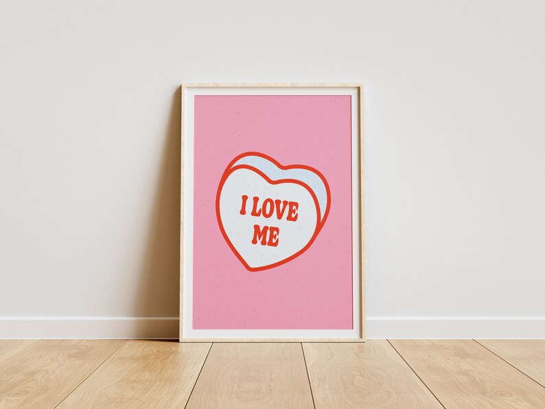 A framed retro-inspired poster featuring two overlapping blue-colored candy hearts with the words 'I Love Me' in bold red text. The design is set against a soft pink background. Displayed against a white wall.