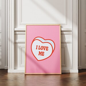 A framed retro-inspired poster featuring two overlapping blue-colored candy hearts with the words 'I Love Me' in bold red text. The design is set against a soft pink background. Displayed against a white wall.