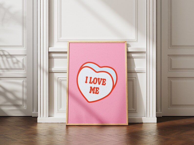 A framed retro-inspired poster featuring two overlapping blue-colored candy hearts with the words 'I Love Me' in bold red text. The design is set against a soft pink background. Displayed against a white wall.
