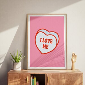 A framed retro-inspired poster featuring two overlapping blue-colored candy hearts with the words 'I Love Me' in bold red text. The design is set against a soft pink background. Displayed on a beautiful sideboard cabinet.