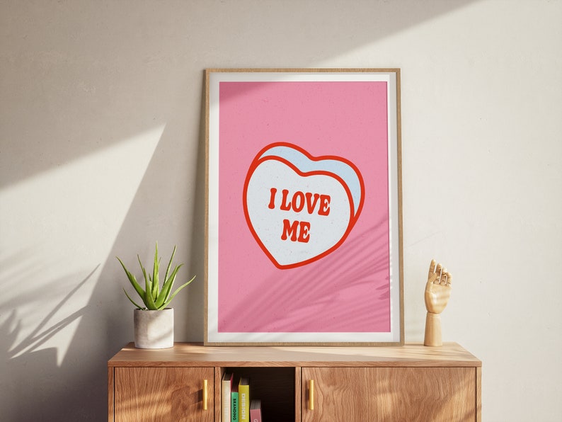 A framed retro-inspired poster featuring two overlapping blue-colored candy hearts with the words 'I Love Me' in bold red text. The design is set against a soft pink background. Displayed on a beautiful sideboard cabinet.