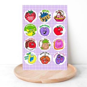 May include: A sheet of colorful fruit-themed stickers with cartoon illustrations and text. The stickers feature a strawberry, blueberries, banana split, banana, plum, apple, pineapple, grapes, pear, and more. The background is a purple and white checkered pattern.