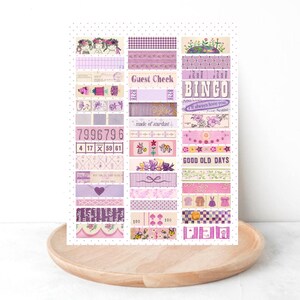 Peut inclure: Une feuille de bandes de ruban adhésif décoratif washi dans les tons violets et roses. Les bandes présentent divers motifs, notamment des motifs floraux, du vichy et du texte tel que "Guest Check" et "BINGO". La feuille est présentée sur un plateau en bois.