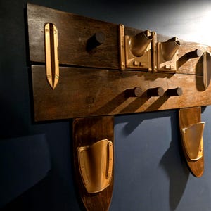 May include: A wall-mounted coat rack made of two horizontal dark brown wooden planks. It features five dark wooden pegs, two gold-toned metal wall pockets at the top, two gold-toned metal wall pockets at the bottom, and two gold-toned metal accents.