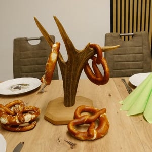 May include: A decorative antler centrepiece with pretzels hanging from the tines. Several pretzels are on the wooden table. A plate and green napkin are also on the table. The antler is mounted on a wooden base.