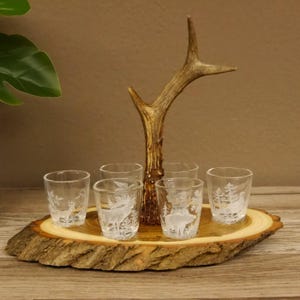Könnte beinhalten: Ein Set aus sechs Schnapsgläsern mit geätzten Waldszenen, darunter Hirsche und Bäume. Die Gläser stehen auf einem Holztablett mit natürlicher Kante und einem dekorativen Geweih.