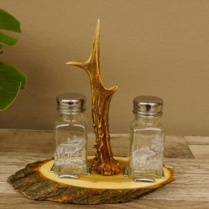 Könnte beinhalten: Ein rustikales Salz- und Pfefferstreuer-Set. Das Set enthält zwei klare Glasstreuer mit geätzten Wildtierdesigns, einem Hirsch und einem Wildschwein, und ein natürliches Geweih-Mittelstück. Die Streuer stehen auf einem Holzsockel.