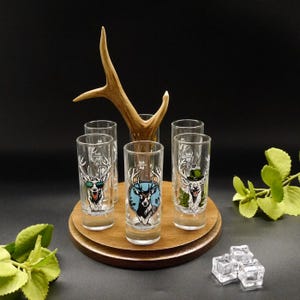 Op de afbeelding: Zes heldere shotglaasjes met hertenillustraties, elk met een uniek ontwerp, zijn gerangschikt op een houten voet. Een hertengewei is achter de glazen geplaatst. De glazen zijn versierd met kleurrijke herten gezichten.