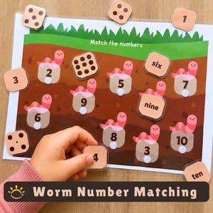 May include: A laminated educational activity sheet titled Match the numbers featuring a brown soil background with green grass and pink worm illustrations. The sheet includes various square tiles with numbers and words to match with the worms.
