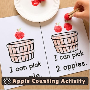 May include: A white paper worksheet featuring two illustrations of brown baskets with red pom-pom apples. The text reads I can pick apple and I can pick 2 apples. A wooden clothespin holds a red pom-pom above a white paper plate with red paint.