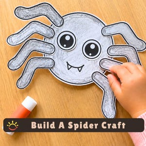 May include: A paper spider craft featuring a round gray body with large black and white eyes and two small fangs. The spider has eight gray legs attached to the body. A glue stick is visible nearby. The text Build A Spider Craft is present.