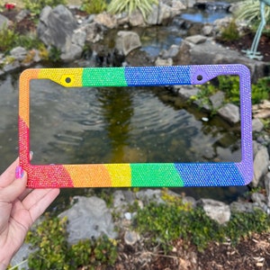 May include: A rainbow-colored license plate frame with rhinestones. The frame is made of metal and has a shiny finish. The colors of the rainbow are red, orange, yellow, green, blue, indigo, and violet.