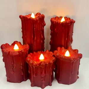 Puede incluir: Cinco velas LED rojas con apariencia de cera derretida. Las velas tienen una superficie texturizada y una luz blanca cálida parpadeante. Las velas son de diferentes alturas y están dispuestas juntas.
