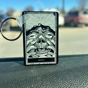 May include: A black and white keychain featuring a tarot card design. The card depicts a detailed illustration of a face with the word "DEATH" at the bottom. The keychain has a clear plastic loop and a metal ring.
