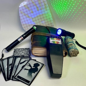 Könnte beinhalten: Eine Sammlung von paranormalen Untersuchungswerkzeugen, darunter ein schwarzer Laserpointer, ein ledergebundenes Notizbuch, ein EMF-Messgerät, ein Salbeibündel, ein Tarotkartenspiel und eine schwarze Taschenlampe. Die Tarotkarten zeigen Schwarz-Weiß-Illustrationen.