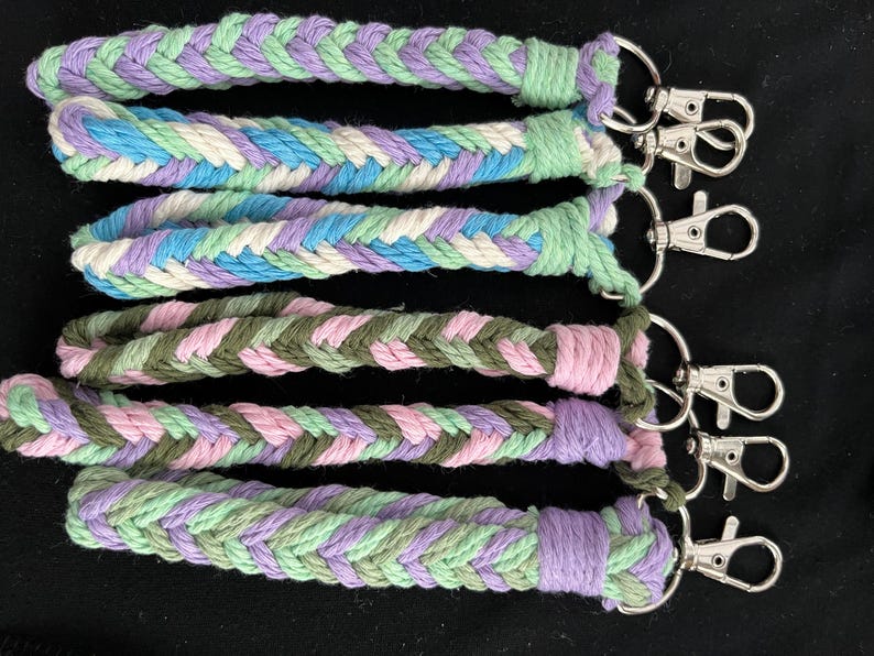 May include: A selection of braided rope keychains in pastel shades of lavender, blue, green, and pink. Each keychain features a silver-coloured clasp for attaching keys. The keychains are made from thick, woven rope.