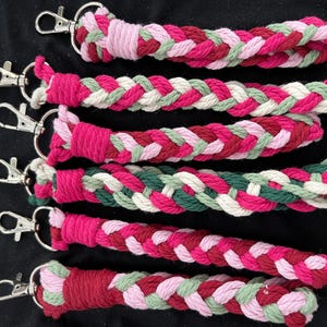 May include: A collection of braided keyrings in shades of pink, red, green, and white. Each keyring has a silver-coloured clasp. The keyrings are made from a soft, woven material.