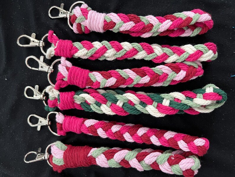 May include: A collection of braided keyrings in shades of pink, red, green, and white. Each keyring has a silver-coloured clasp. The keyrings are made from a soft, woven material.
