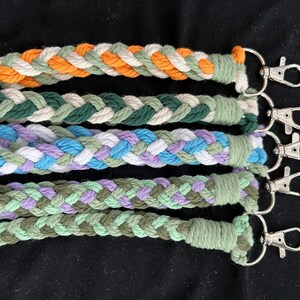 May include: Five braided keychains in various colours: orange, green, blue, purple, and olive. Each keychain has a silver-coloured clasp. The keychains are made of a thick, woven material.