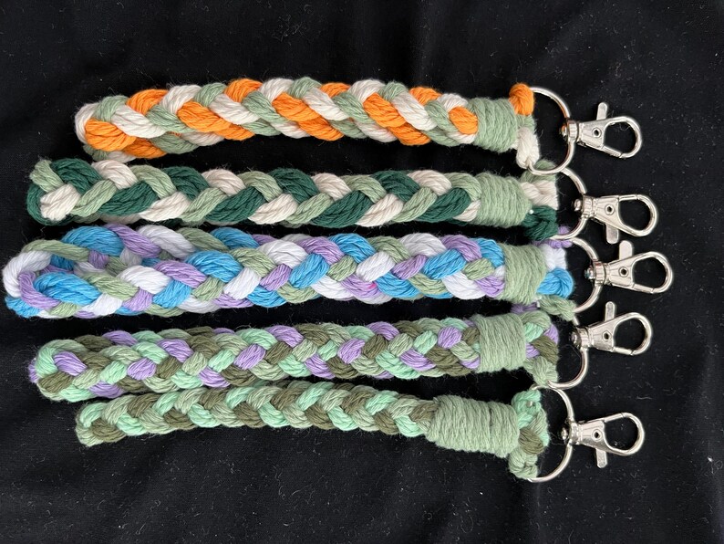May include: Five braided keychains in various colours: orange, green, blue, purple, and olive. Each keychain has a silver-coloured clasp. The keychains are made of a thick, woven material.