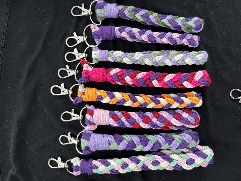 May include: Braided keyrings in a variety of colours, including purple, pink, orange, and green. Each keyring has a silver-coloured clasp. The keyrings are displayed on a black surface.