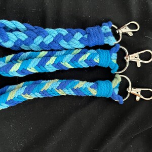 May include: Three braided keychains in shades of blue and green. Each keychain features a different pattern of woven cotton cord and a silver-coloured clasp. The keychains are displayed on a black surface.
