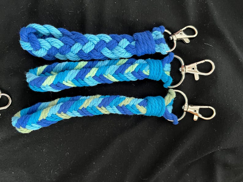 May include: Three braided keychains in shades of blue and green. Each keychain features a different pattern of woven cotton cord and a silver-coloured clasp. The keychains are displayed on a black surface.