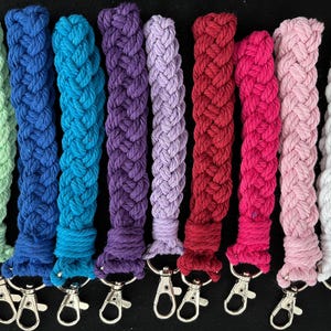 May include: A selection of braided rope keychains in a range of colours: green, blue, purple, red, pink, and white. Each keychain has a silver-coloured clasp for easy attachment. The keychains are shown against a black background.