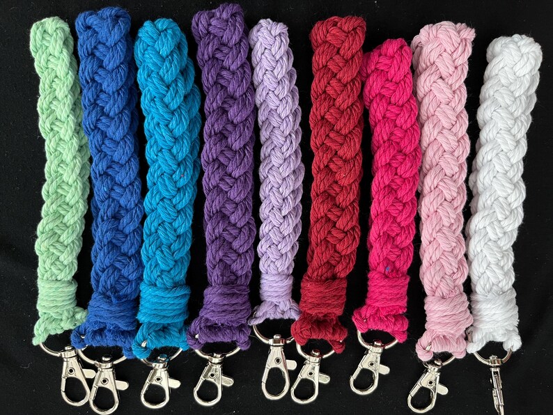 May include: A selection of braided rope keychains in a range of colours: green, blue, purple, red, pink, and white. Each keychain has a silver-coloured clasp for easy attachment. The keychains are shown against a black background.