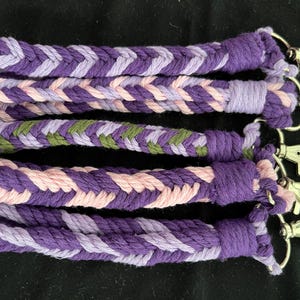 May include: Braided wrist straps in shades of purple, pink, and green, each with a silver-coloured clasp. The straps are made of woven cotton and are approximately 18 cm long.