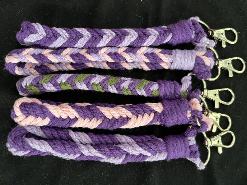 May include: Braided wrist straps in shades of purple, pink, and green, each with a silver-coloured clasp. The straps are made of woven cotton and are approximately 18 cm long.