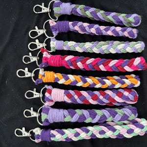 May include: Braided keyrings in a variety of colours, including purple, pink, orange, and green. Each keyring has a silver-coloured clasp. The keyrings are displayed on a black surface.