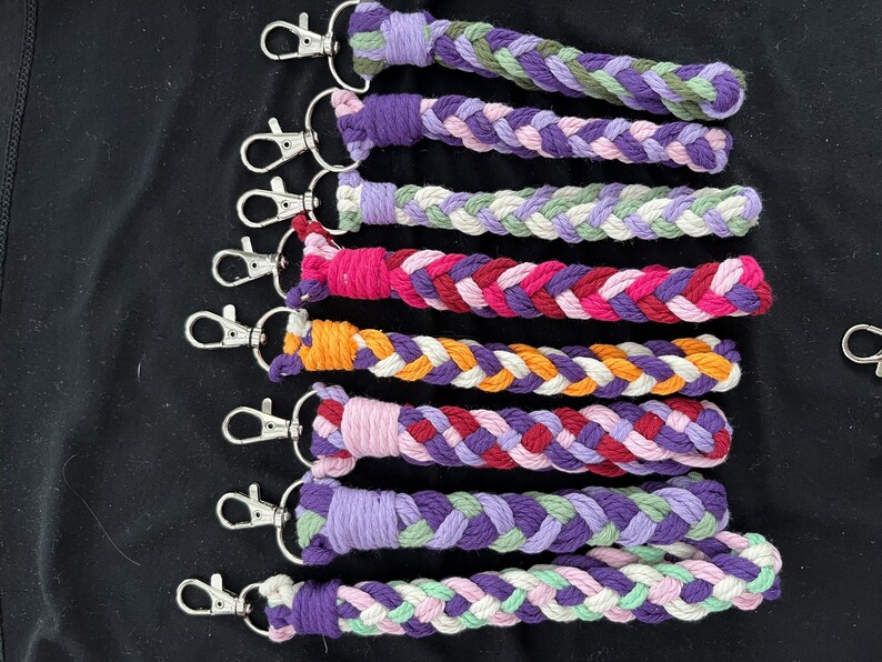 May include: Braided keyrings in a variety of colours, including purple, pink, orange, and green. Each keyring has a silver-coloured clasp. The keyrings are displayed on a black surface.